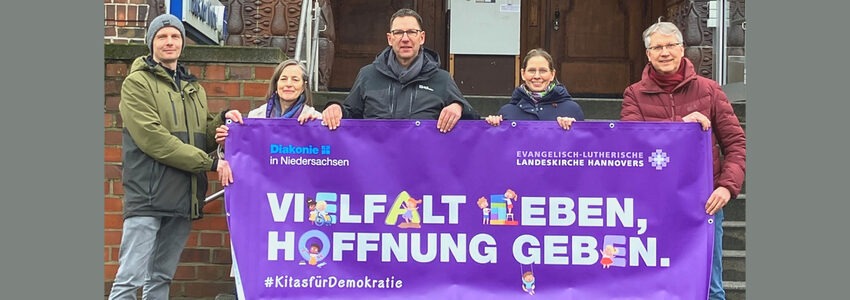 Foto: Ev.-luth. Kindertagesstättenverband Lüneburg. Das Transparent mit dem Motto der Kampagne halten von links: Matthias Willimzig (Stv. Kitaleitung St. Johannis), Friederike Orth (päd. Geschäftsführung), Christian Stasch (Superintendent), Dorothea Noordveld-Lorenz (Vorstandsvorsitzende des Ev.-luth. Kitaverbandes), Christian Cordes (Superintendent).