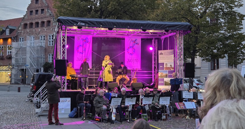 Foto: Lüne-Blog. "Deutschland singt" - 3. Oktober 2025. OB Claudia Kalisch begrüßt die Menschen auf dem Marktplatz. Im Vordergrund:  Posaunenchor des Kirchenkreises Lüneburg. Rechts auf der Bühne Klaus Stehr von Noordlücht.