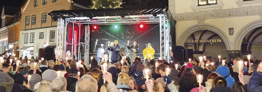 Foto: T. Hüske. Weihnachtsliedersingen vor dem Rathaus in Lüneburg. OB Claudia Kalisch steht am Mikro auf der Bühne, die Zuhörenden halten brennende Kerzen in die Höhe.
