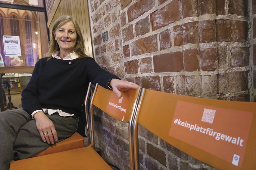 Foto: T. Hüske.  Pastorin Silke Ideker, St. Michaelis, Lüneburg, auf den drei orangefarbenen Stühlen, die im Kirchenraum aufgestellt sind. Sie sollen zeigen, "dass wir keinen Platz für Gewalt gegen Frauen haben. Wir wollen dafür eintreten, dass hingesehen wird."