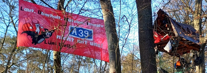 Foto: Cyclocracy. „Laut. Überholt. Nutzlos. Invasiv.“ - So die kurz gefasste Stellungnahme auf dem roten Transparent im Lüner Holz zu den Bauplänen für die A39. Rechts daneben Plattform und Baumhaus.