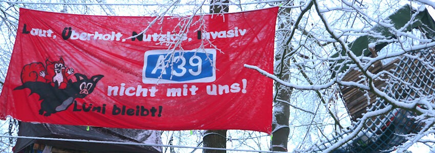 Foto: Gruppe Flederhörnchen. Im verschneiten Lüner Holz hängt neben den beiden Baumhäusern das Transparent "Lüni bleibt".