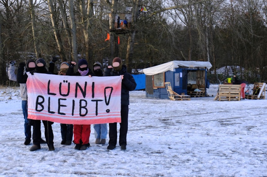 Foto: Initiative "Voßbrook für alle!". Die Gruppe hält seit Oktober 2025 eine Plattform auf Bäumen auf dem MFG5-Gelände in Kiel-Holtenau besetzt. Sie protestieren mit der Aktion gegen den drohenden Verkauf des Geländes durch die Stadt Kiel an die Bundeswehr. Sie setzen sich dafür ein, dass das Gelände als allen Menschen zugänglicher und vielfältiger Ort erhalten und entsprechend gestaltet wird.