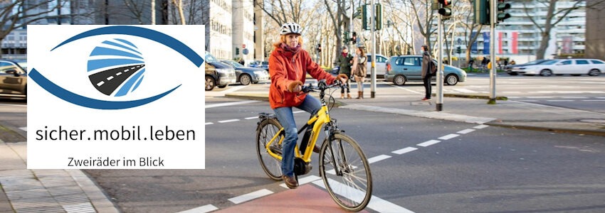 Foto: Rendel-Freude (DVW) mit Logo „sicher.mobil.leben“ (Polizei Hamburg). Frau auf einem E-Bike an einer Kreuzung.