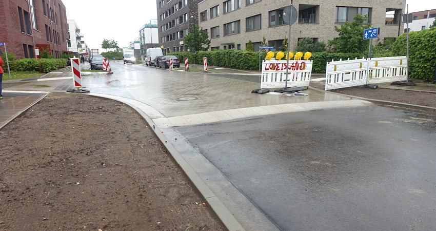 Foto: Uwe Wenk, 11.05.2026. Ort: Lübecker Straße. Links geht es in die Marga-Jess-Straße, rechts in die Hans-Heinricht-Stelljes-Straße. Positiv: Signalfelder in der Längsrichtung zur Straße weisen auf die Querung hin. Der Gehweg ist hier auch plan abgesenkt. Die sog. Sinussteine und die Aufpflasterung signalisieren dem Kfz-Verkehr: Vorsicht, Querungstelle!
Negativ: Warum gibt es hier keine Querungsmöglichkeit für Fußgänger, die von rechts nach links über die Straße gehen wollen?